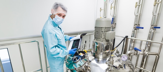 Científico de imágenes panorámicas con traje de laboratorio azul y máscara de gas en una sala limpia y revisa los gráficos en una tableta cerca de un gran tanque de acero