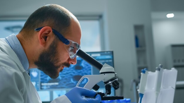 Científico de investigación hombre mirando muestras bajo microscopio en laboratorio equipado moderno