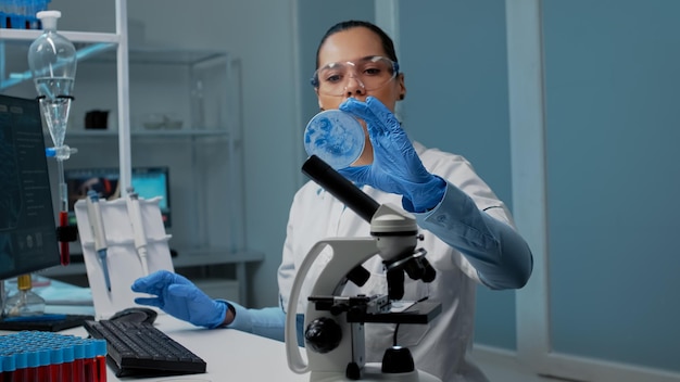Foto gratuita científico químico con equipo de investigación en laboratorio en escritorio con placa de petri, micropipeta, vacutainers y microscopio. mujer científica escribiendo en computadora para el desarrollo