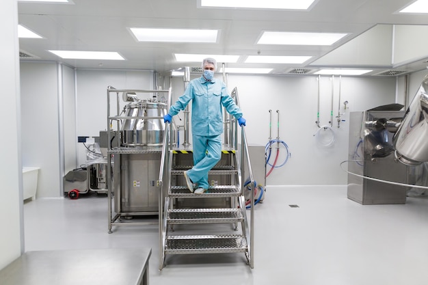 Científico en traje de laboratorio azul parado en escaleras de metal cromado con piernas cruzadas