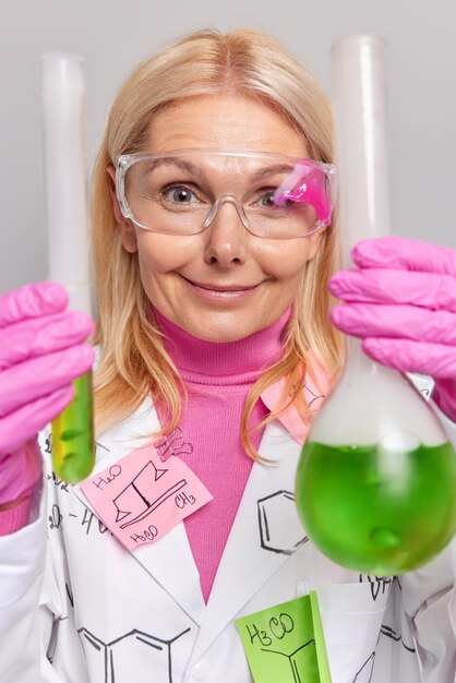 Científico en uniforme sostiene dos matraces con reactivos verdes demuestra reacciones químicas visuales usa anteojos protectores aislados en gris