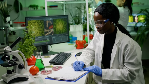 Científicos investigadores que trabajan en el laboratorio de biotecnología mirando comida vegana y árboles jóvenes analizando mutaciones genéticas escribiendo experiencia biológica en un bloc de notas. Bioquímica de investigación farmacéutica