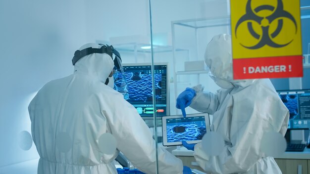 Científicos en mono de pie detrás de la pared de vidrio que trabajan en la zona de peligro del laboratorio