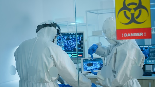 Científicos en mono de pie detrás de la pared de vidrio que trabajan en la zona de peligro del laboratorio