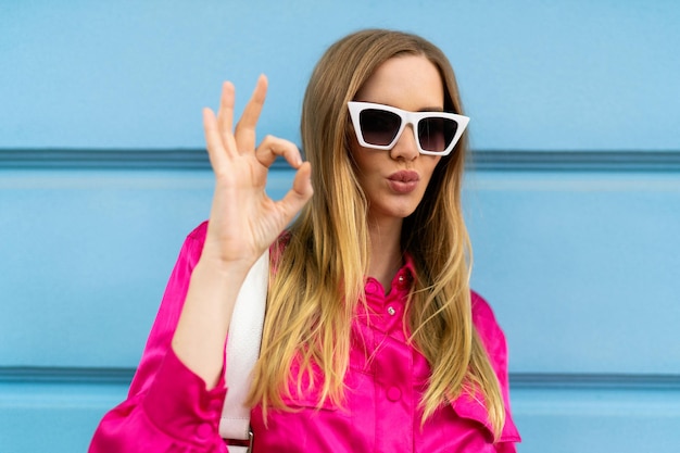 Cierra el brillante retrato positivo de una mujer rubia influyente bloguera de moda, vestida con un traje brillante y gafas de sol posando cerca de la pared azul.