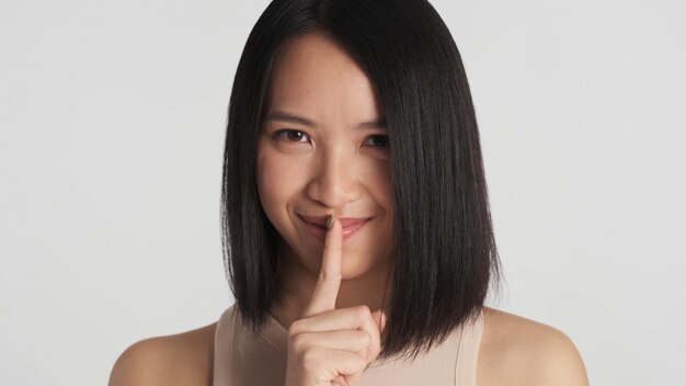 Cierra a una chica asiática que se ve feliz mostrando un gesto de silencio a la cámara sobre fondo blanco Mantener secreto