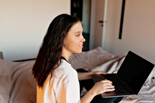 Cierra la foto interior de una chica de cabello oscuro con camisa blanca trabajando en casa con una laptop en la cama