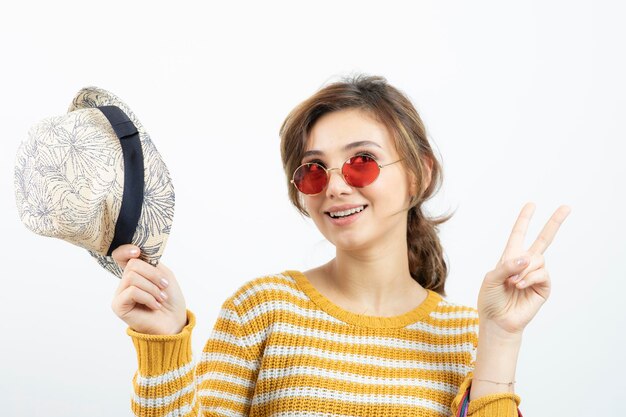 Cierra la foto de una mujer hermosa con sombrero y gafas mostrando el signo de la victoria. foto de alta calidad