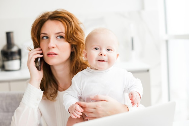Cierra la foto de una mujer de negocios sentada y hablando por su celular mientras sostiene a su pequeño bebé sonriente en la mano