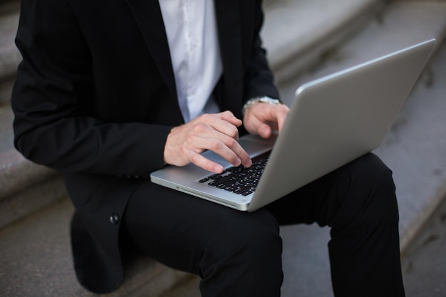 Cierra a un hombre de negocios con traje negro clásico trabajando en una laptop al aire libre