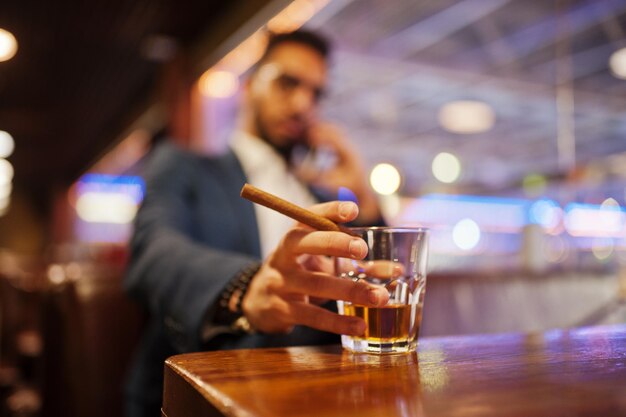 Cierra la mano de un apuesto hombre árabe bien vestido con un vaso de whisky y un cigarro posado en el pub