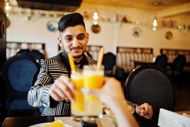 Cierra las manos de una encantadora pareja india enamorada sentada en el restaurante y animando juntos con jugo de naranja