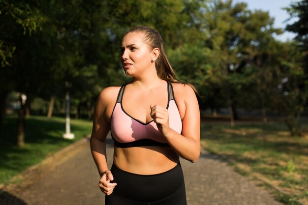 Cierra a una mujer exhausta de talla grande con top deportivo y calzas mirando cansadamente a un lado mientras corres en el parque de la ciudad