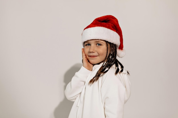 Cierra el retrato de una adorable y encantadora niñita con coletas con gorra de Papá Noel y vestido blanco sonriendo y posando ante la cámara sobre un fondo aislado