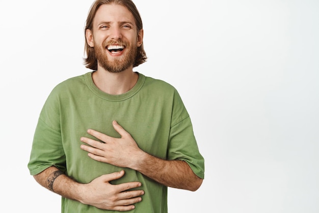 Cierra el retrato de un apuesto hombre rubio, riendo y sonriendo feliz, expresión facial despreocupada, estado de ánimo positivo, de pie con una camiseta verde sobre fondo blanco.