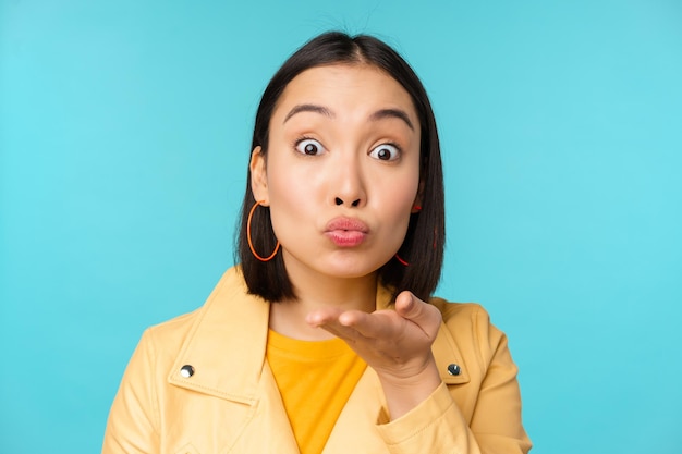 Cierra el retrato de una chica asiática divertida que envía un beso de aire que sopla a la cámara con los ojos reventados de pie sobre un fondo azul.