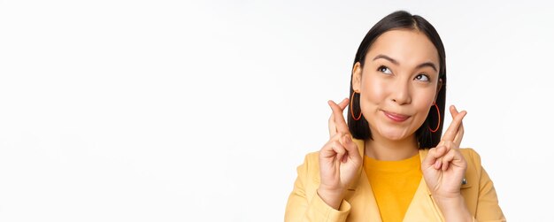 Cierra el retrato de una chica asiática esperanzada deseando cruzar los dedos para tener buena suerte rezando y sonriendo de pie sobre fondo blanco.