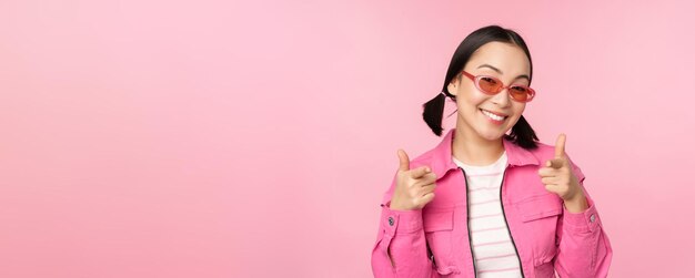 Cierra el retrato de una chica asiática moderna con gafas de sol sonriendo señalando con el dedo a la cámara elogiándote invitando o halagando de pie sobre un fondo rosa