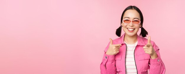 Cierra el retrato de una chica asiática moderna con gafas de sol sonriendo señalando con el dedo a la cámara elogiándote invitando o halagando de pie sobre un fondo rosa