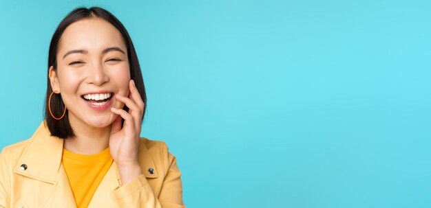 Cierra el retrato de una chica asiática natural riéndose sonriendo y luciendo feliz de pie sobre fondo azul.