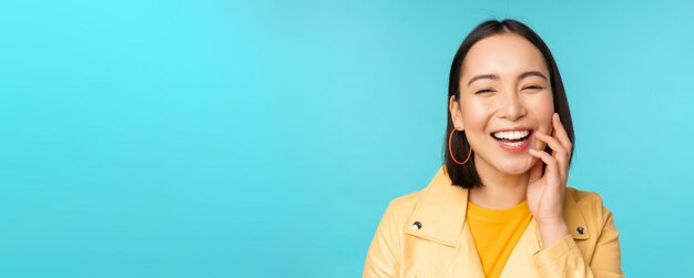 Cierra el retrato de una chica asiática natural riéndose sonriendo y luciendo feliz de pie sobre fondo azul.