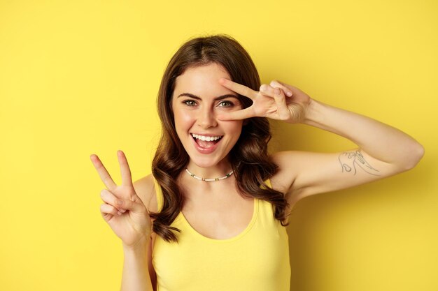 Cierra el retrato de una chica feliz con estilo, mostrando el signo de la paz y sonriendo, posando contra un fondo amarillo