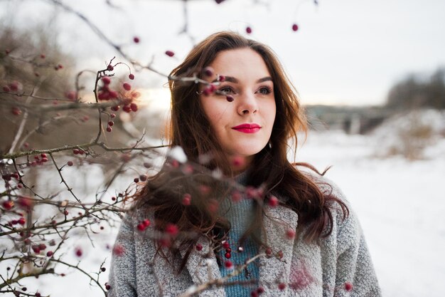 Cierra el retrato de una chica gentil con abrigo gris cerca de las ramas de un árbol cubierto de nieve