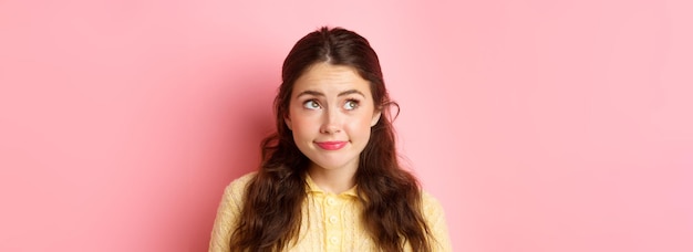 Foto gratuita cierra el retrato de una chica linda y modesta sonriendo y mirando tímidamente en la esquina superior izquierda de pie pensativo usando maquillaje de fondo rosa