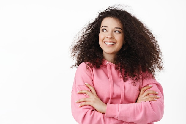 Cierra el retrato de una chica moderna y elegante con el pelo rizado, con capucha rosa, brazos cruzados en el pecho, sonriendo y mirando a un lado el logo, de pie sobre fondo blanco.