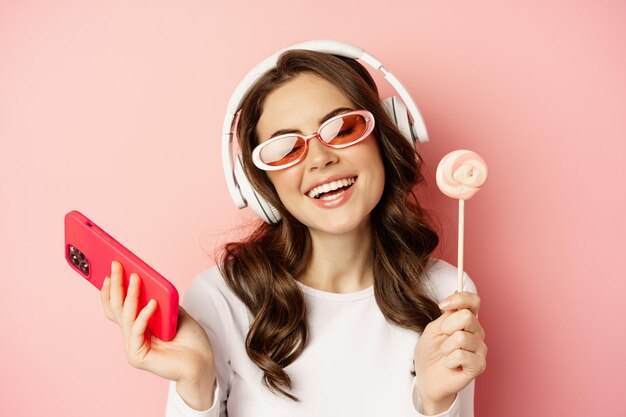 Cierra el retrato de una elegante chica glamurosa, una mujer hermosa con auriculares, escuchando música con un smartphone, usando gafas de sol y lamiendo lolipop.