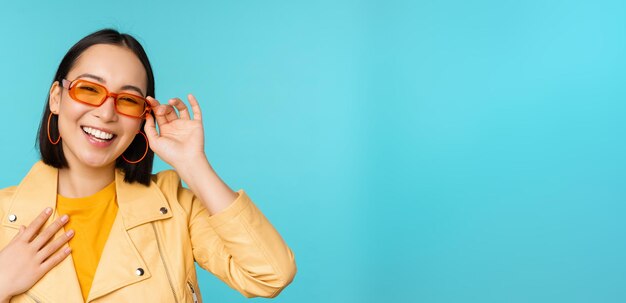 Cierra el retrato de una elegante mujer asiática con gafas de sol riendo y sonriendo feliz posando con ropa de moda sobre fondo azul.