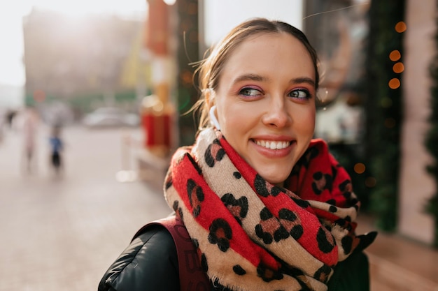 Cierra el retrato exterior de una mujer feliz sonriente con una sonrisa perfecta y un maquillaje brillante usando una bufanda brillante mirando a un lado y sonriendo mientras camina por la ciudad para reunirse