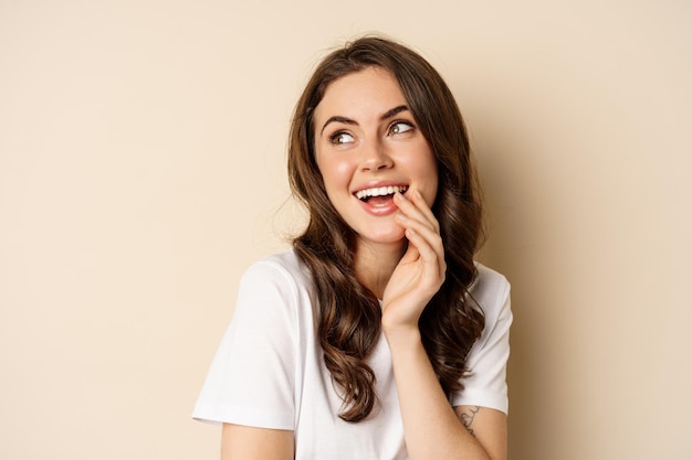 Cierra el retrato de una hermosa chica coqueta que mira tontamente, sonríe suavemente y se ríe, posando coqueta y linda contra un fondo beige. Concepto de belleza y san valentín de las mujeres.