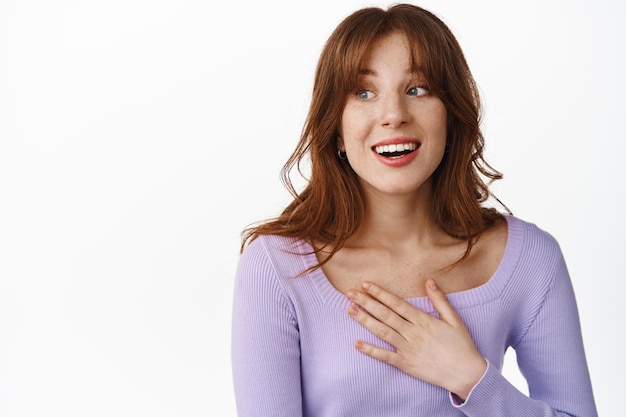 Cierra el retrato de una hermosa chica con estilo, con pecas y maquillaje, con la mano en el corazón, sonriendo y mirando sorprendida a la izquierda, mirando la pancarta del logo, de pie sobre fondo blanco