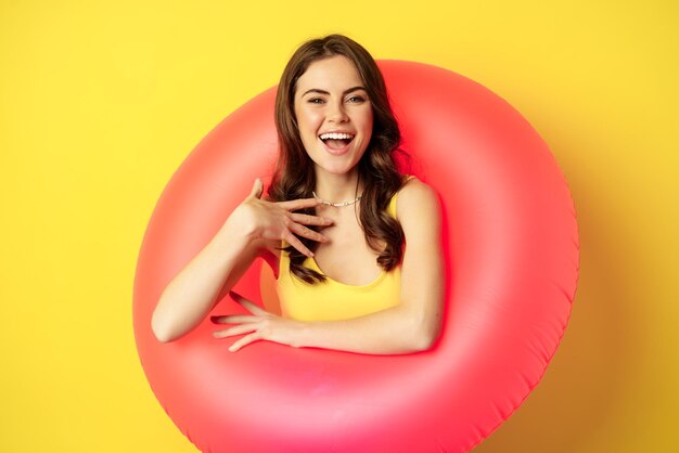 Cierra el retrato de una hermosa modelo de mujer morena dentro de un anillo de natación rosa, accesorios de playa y concepto de vacaciones de verano, fondo amarillo.