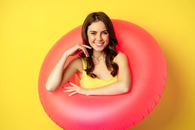 Cierra el retrato de una hermosa modelo de mujer morena dentro de un anillo de natación rosa, accesorios de playa y concepto de vacaciones de verano, fondo amarillo.