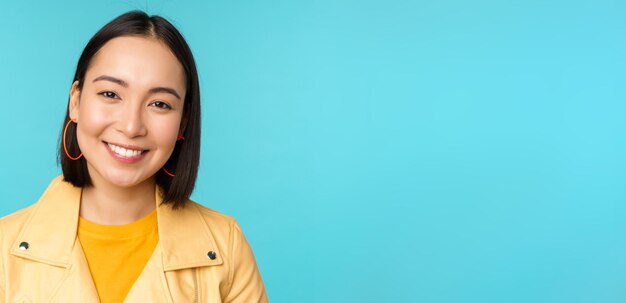 Cierra el retrato de una hermosa mujer asiática sonriente con dientes blancos mirando feliz a la cámara posando con una chaqueta amarilla sobre el fondo azul del estudio