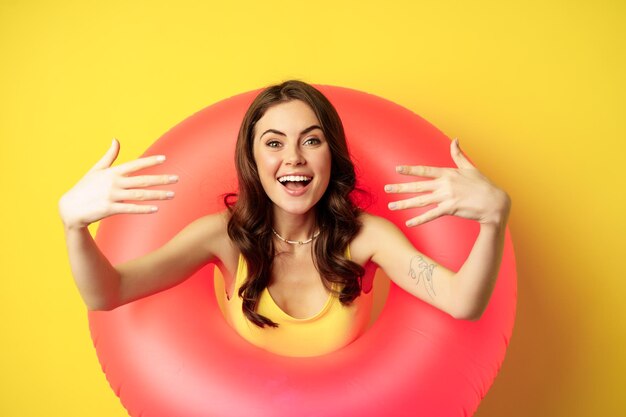 Cierra el retrato de una hermosa mujer feliz dentro del anillo de natación rosa, atrayendo, invitando a la gente a las vacaciones de verano, a la fiesta en la playa, de pie sobre un fondo amarillo.