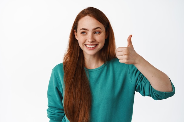 Cierra el retrato de una hermosa mujer joven con el pelo largo rojo, muestra el pulgar hacia arriba y sonríe complacido, mirando el texto promocional del logotipo a un lado, de pie sobre un fondo blanco.
