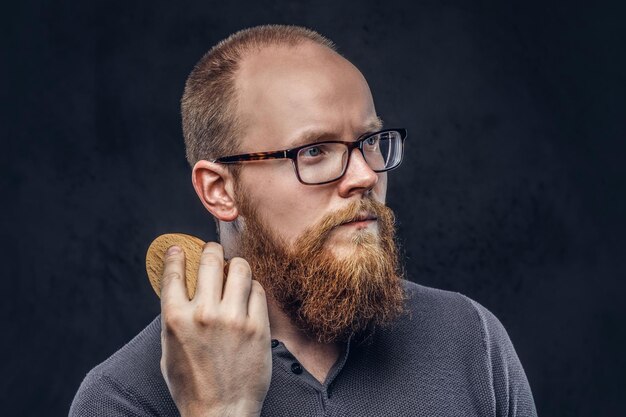 Cierra el retrato de un hombre barbudo pelirrojo que usa anteojos vestido con una camiseta gris, se preocupa por su barba usando un cepillo de barba. Aislado en un fondo de textura oscura.