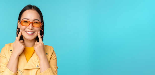 Cierra el retrato de una joven asiática con gafas de sol sonriendo y luciendo romántica de pie feliz sobre fondo azul.