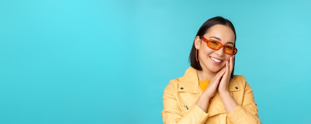 Cierra el retrato de una joven asiática con gafas de sol sonriendo y luciendo romántica de pie feliz sobre fondo azul.