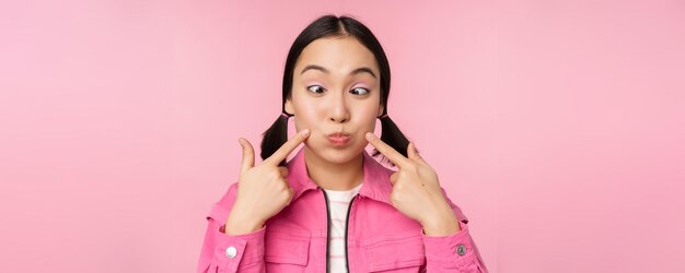 Cierra el retrato de una joven asiática mostrando sus hoyuelos metiendo las mejillas tontamente y haciendo muecas de pie sobre un fondo rosa