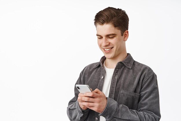Cierra el retrato de un joven, estudiante universitario enviando mensajes de texto por teléfono, chatea con un amigo en la aplicación de redes sociales para teléfonos inteligentes, usa la aplicación, parado sobre fondo blanco
