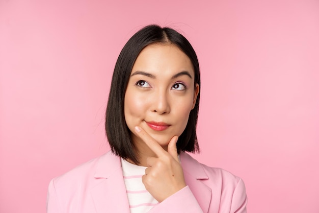 Cierra el retrato de una joven mujer de negocios asiática pensando sonriendo pensativa y mirando la esquina superior izquierda de pie sobre un fondo rosa