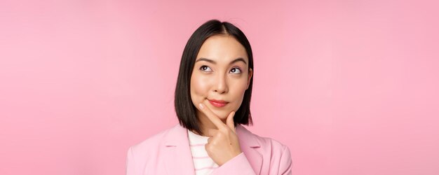 Cierra el retrato de una joven mujer de negocios asiática pensando sonriendo pensativa y mirando la esquina superior izquierda de pie sobre un fondo rosa