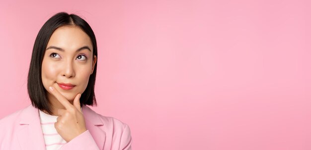 Cierra el retrato de una joven mujer de negocios asiática pensando sonriendo pensativa y mirando la esquina superior izquierda de pie sobre un fondo rosa