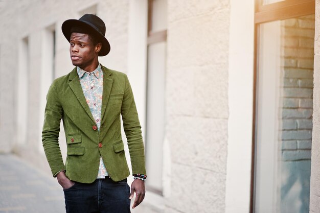 Cierra el retrato de moda de un hombre afroamericano negro con una chaqueta de terciopelo verde y un sombrero negro caminando por las calles de la casa de fondo de la ciudad con muchas ventanas