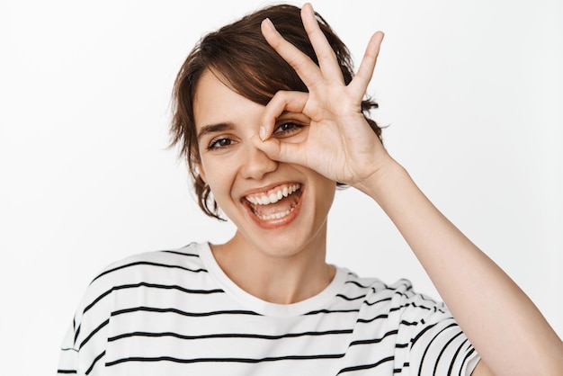 Cierra el retrato de una mujer feliz sonriente que muestra el signo de ok y parece satisfecha recomendando algo bueno elogiando el producto de pie sobre fondo blanco.