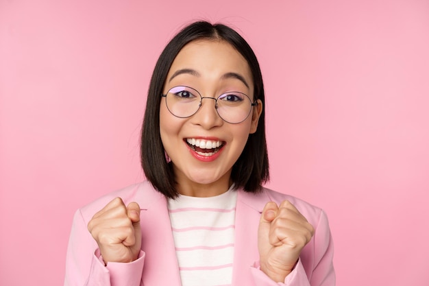 Cierra el retrato de una mujer de negocios asiática animando a enraizar por mirar con esperanza y emoción a la cámara sonriendo y riendo de pie sobre un fondo rosa